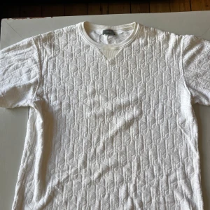 Vit t-shirt med mönster från Dior - Säljer en äkta vit t-shirt från Dior med upphöjt, diskret mönster över hela plagget. T-shirten är i 100% bomull och har rund halsringning och korta ärmar. Perfekt för dig som gillar exklusiva och stilrena plagg. Köpt från vestiaire för 6 000 kr 