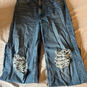 Blå slitna wide jeans från H&M Divided - Säljer ett par blå jeans från H&M Divided med breda ben och slitna detaljer på båda knäna. Jeansen har klassisk femficksdesign och hög midja. Perfekta för dig som gillar en avslappnad och trendig stil.