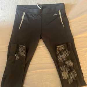 Svarta träningstights från H&M Sport - Svarta träningstights från H&M Sport med gråa detaljer och mönster på benen. Modellen är trekvarts och har praktiska fickor på sidorna. Perfekta för gym eller löpning.