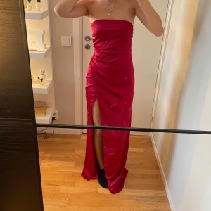 Röd axelbandslös balklänning från Nelly - Säljer en röd axelbandslös långklänning från Nelly med slits framtill. Klänningen har en figurnära passform och är tillverkad i ett glansigt material som ger en lyxig känsla. Perfekt för speciella tillfällen.