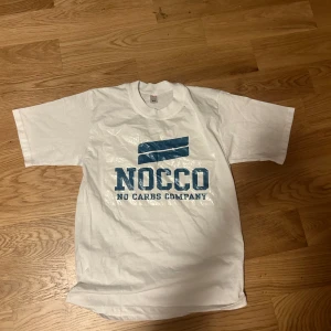 Vit NOCCO t-shirt med blå logga - Vit t-shirt från NOCCO med stor blå logga och texten 'NOCCO NO CARBS COMPANY' på bröstet. Klassisk passform och rund halsringning. Perfekt för dig som gillar sportiga och enkla plagg.