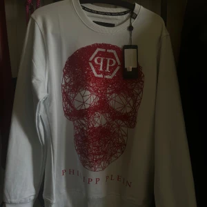 Vit sweatshirt med röd döskalle från Philipp Plein - Säljer en ny vit sweatshirt från Philipp Plein med en stor röd glittrig döskalle framtill och logga på bröstet. Tröjan har rund halsringning och långa ärmar. Skriv för frågor och bilder