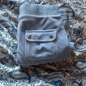 Ljusblå mjukisbyxor från Juicy Couture - Säljer ett par ljusblå mjukisbyxor från Juicy Couture med bred resår i midjan och snörning. Byxorna har en ficka med lock och tryckknappar på ena benet. Perfekta för en avslappnad stil.