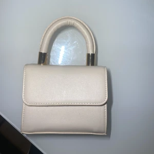 Beige mini axelväska med gulddetaljer - Säljer en beige mini axelväska med stilrena gulddetaljer på handtaget. Väskan har ett avtagbart axelband och ett klassiskt, stilrent utseende. Perfekt för dig som gillar minimalistisk design och vill ha en liten väska till det viktigaste. Perfekt till sommaren! ☀️🥂