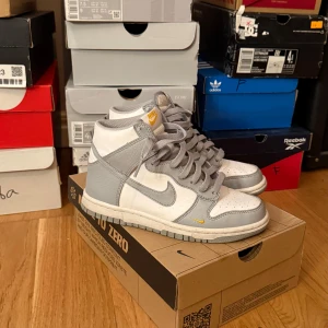 Nike Dunk High NN GS grå och vita sneakers - Nike Dunk High NN GS sneakers i vitt och ljusgrått med gula detaljer. Perfekta för dig som gillar streetwear och sneakers. Säljer då jag inte använder dem de är använda få tal gånger och är i väldigt bra skick. Hör av dig vid funderingar eller prisförslag 💞