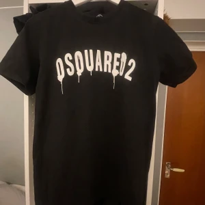 Svart t-shirt från Dsquared2 med tryck - Säljer en svart t-shirt från Dsquared2 i storlek M. T-shirten har ett vitt tryck med texten 'DSQUARED2' på bröstet där bokstäverna har en droppande effekt. Klassisk passform och rund hals.