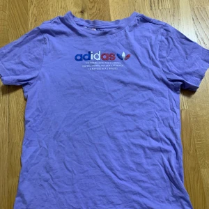 Lila t-shirt från Adidas - Lila t-shirt från Adidas med tryckt logga i blått, vitt och rött framtill. Klassisk rund halsringning och korta ärmar. Perfekt för en sportig och avslappnad stil.