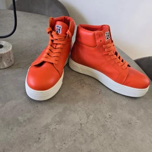 Orange sneakers från Tommy Hilfiger - Snygga orange sneakers från Tommy Hilfiger med hög topp och snörning. Perfekta för en sportig look med en touch av färg. I läder, använda en gång.