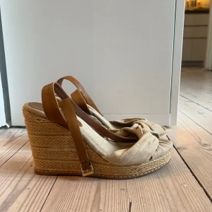 Beige kilklackar från Tommy Hilfiger - Så snygga och bekväma beige kilklackar från Tommy Hilfiger med bruna remmar och knutdetalj framtill. Så snygg till jeans eller vit klänning till studenten. De har tecken på användning, storlek 37-38 (liten 38) 