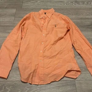 Persikofärgad button-down oxfordskjorta skjorta från Polo Ralph Lauren - Säljer en persikofärgad långärmad button-down oxfordskjorta från Polo Ralph Lauren i slim fit. Skjortan har klassisk krage, knappar framtill och broderad logga på bröstet. Tillverkad i bomull med stretch för extra komfort. Ny pris på denna skjortan ligger på 1600kr