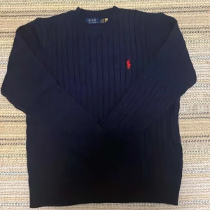 Ralph lauren ull tröja  - Klassisk mörkblå kabelstickad tröja från Polo Ralph Lauren med röd broderad logga på bröstet. Tröjan har lång ärm och rund halsringning, perfekt för en stilren och tidlös look.