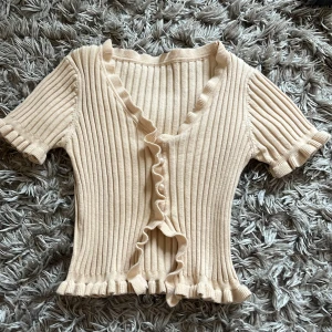 Beige ribbad topp med volangkant - Superfin beige ribbad topp med korta ärmar och volangdetaljer längs v-ringningen och nederkanten. Perfekt att styla till jeans eller kjol för en trendig look.