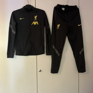 Svart träningsset från Nike x Liverpool FC - Säljer ett svart träningsset från Nike i samarbete med Liverpool FC. Setet består av en zip-jacka och matchande byxor med gula loggor och detaljer. Storlek på tröjan är 158-170 och storleken på byxorna är 170