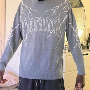 Gråblå stickad tröja med blixtmönster och text - Säljer en gråblå stickad tröja med vita blixtar och stor text. Tröjan har rund halsringning och långa ärmar. Perfekt för dig som gillar streetwear och vill sticka ut med ett coolt mönster.