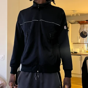 Svart track jacket från Moncler - Säljer en svart track jacket från Moncler med dragkedja framtill och hög krage. Jackan har en vit dekorlinje över bröstet och axlarna samt Moncler-logga på ärmen. 