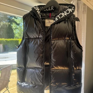 Svart dunväst från Moncler med huva - Säljer en svart glansig dunväst från Moncler med huva och dragkedja. Västen har två fickor med dragkedja framtill och Moncler-logga på ryggen. Perfekt för lager-på-lager och snygg streetstil. Inte äkta! Passar L