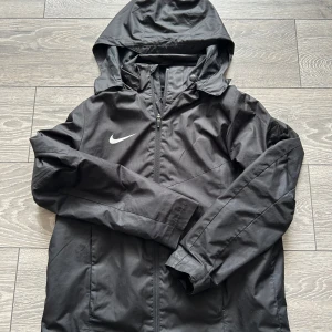 Svart vindjacka från Nike med huva - Säljer en svart vindjacka från Nike med huva och dragkedja framtill. Jackan har lång ärm, diskret Nike-logga på bröstet och är i loose fit. Perfekt för blåsiga dagar.