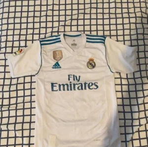 Real Madrid hemmatröja - Säljer en vit Real Madrid hemmatröja från Adidas med blå detaljer och klubbmärke på bröstet. Tröjan har korta ärmar och Fly Emirates som sponsortryck på framsidan. Perfekt för fotbollsfans.Ronaldo på bakisdan också!   Start pris 90 pris kan diskuteras också!