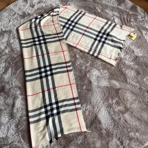 Beige rutig halsduk - Säljer en klassisk beige halsduk med det ikoniska rutmönstret i svart, vitt och rött. Halsduken har fransar i kanterna och är tunn och mjuk, perfekt för att lyfta vilken outfit som helst.