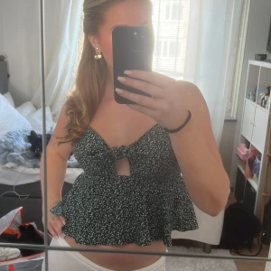 Grön blommig topp med knyt - Säljer en söt grön blommig topp med knyt framtill. Toppen är ärmlös och har en volangdetalj nedtill. Perfekt för varma sommardagar!