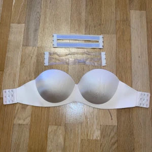 Beige axelbandslös bh - Säljer en beige axelbandslös bh med slät yta och vadderade kupor. Perfekt för klänningar och toppar utan axelband. Inkluderar två olika band, ett transparent och ett i vitt tyg, för extra stöd.