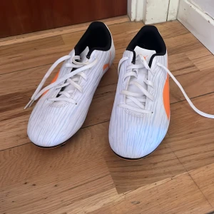 Vita och orange fotbollsskor från Puma - Snygga fotbollsskor från Puma i vitt med orange detaljer och snörning. Skorna har en mönstrad yta och dobbar för bra grepp på planen. Perfekta för fotbollsträning och matcher.