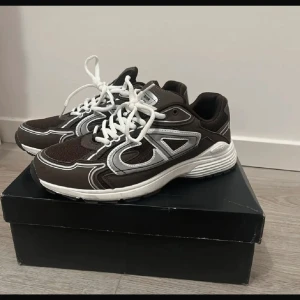 Bruna sneakers från Dior - Snygga bruna sneakers från Dior med vita detaljer och snörning. Skorna har en modern design med en bekväm sula och rund tå. Perfekta för en stilren look. 