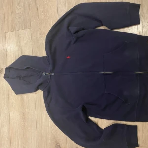 Mörkblå hoodie från Ralph Lauren - Säljer en stilren mörkblå hoodie från Ralph Lauren med dragkedja och en liten röd logotyp på bröstet. Tröjan har långa ärmar och en praktisk huva. Perfekt för en avslappnad stil.