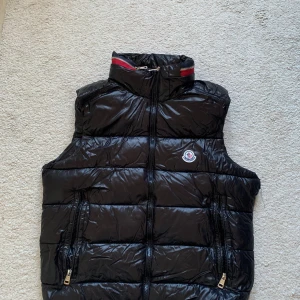 Svart dunväst från Moncler - Snygg svart dunväst från Moncler med dragkedja och hög krage. Perfekt för kyliga dagar när du vill hålla stilen. Västen har två praktiska fickor med dragkedja framtill och Monclers logga på bröstet. !OBS! Västen har två hål vid varje ärm, inget som syns när man har på sig. 