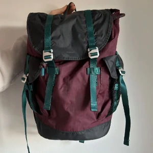 Vinröd och svart ryggsäck med gröna detaljer från Sandqvist - Snygg och praktisk ryggsäck i vinrött och svart med gröna remmar och spännen. Perfekt för vardagsbruk eller utflykter. Ryggsäcken har flera fack och justerbara axelremmar för bekväm passform. Använd enstaka gånger alltså i stort sett nyskick. 