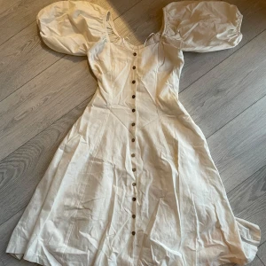 Vit klänning med puffärmar stl xs - Charmig vit klänning med puffiga kortärmar och knappar framtill. Klänningen har en romantisk stil med en lätt och luftig känsla, perfekt för varma dagar. Använd 1 gång