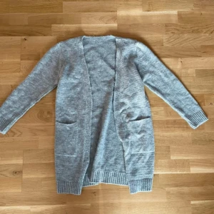 Grå stickad kofta - Mysig grå stickad kofta med långa ärmar och praktiska fickor framtill. Perfekt för kyliga dagar med ribbade muddar vid ärmslut och nederkant. En stilren och bekväm baströja för garderoben.