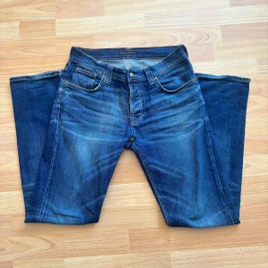Blå jeans från Nudie Jeans - Snygga blå jeans från Nudie Jeans med klassisk femficksdesign och orange sömmar. Jeansen har en normal passform och är tillverkade i slitstarkt denim. Dem är i topp skick och i strorlek 31/32 Perfekta för en avslappnad stil. 