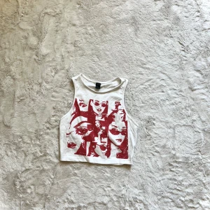 Vit croppad linnetopp med rött tryck - Säljer en vit croppad linnetopp med ett coolt rött tryck av ansikten. Perfekt för en avslappnad stil. Toppen är ärmlös och har en rund halsringning.