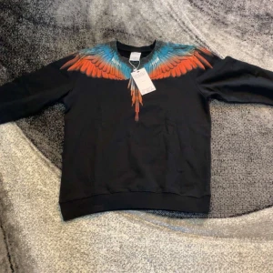  Marcelo Burlon - Säljer en svart tröja från Marcelo Burlon med ett coolt vingmönster i orange och blått över axlarna. Tröjan har långa ärmar och är perfekt för en stilren look. Passar bra för dig som gillar unika designer.