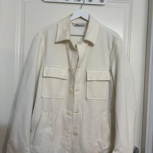 Vit jacka från Zara - Säljer en stilren vit jacka från Zara med knappar framtill och två stora bröstfickor. Jackan har en klassisk krage och är tillverkad i ett mjukt material. Perfekt för en minimalistisk look.