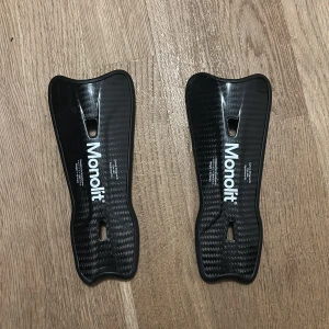 Svarta benskydd från Monolit - Säljer ett par svarta benskydd från Monolit. De har en stilren design med en glansig yta och är tillverkade i ett lätt och hållbart material. Perfekta för fotbollsspelare som söker pålitligt skydd.