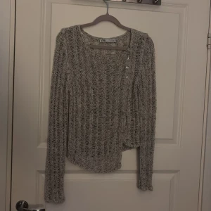 Grå stickad tröja från Zara - Säljer en grå stickad tröja från Zara med en unik design och knappar på sidan. (Går INTE att knäppa upp) Tröjan har en luftig stickning och långa ärmar, perfekt för lager-på-lager-stil. Passar bra till både jeans och kjol. Passar Xs/S/M