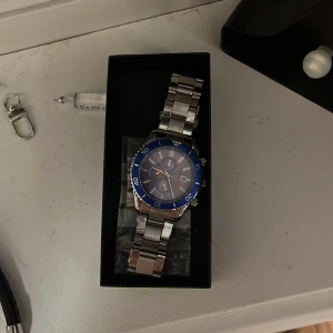 Blå och silverfärgad klocka - Snygg klocka med blå urtavla och silverfärgad metallarmband. Klockan har tydliga markeringar och en vridbar ring runt urtavlan. Perfekt för den som vill ha en stilren accessoar.