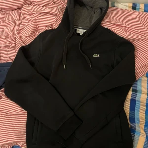 Svart hoodie från Lacoste - Säljer en stilren svart hoodie från Lacoste med grå insida i huvan. Den har en klassisk krokodillogga på bröstet och justerbara snören i huvan. Perfekt för en avslappnad look.