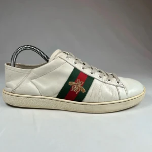 Vita sneakers från Gucci  - Snygga vita sneakers med en broderad bi-detalj på sidan. Skorna har en grön och röd rand som ger en klassisk look. De är tillverkade i skinn och har snörning framtill.