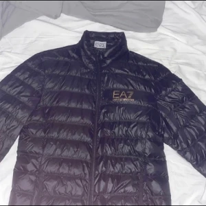 EA7 Emporio Armani dunjacka small - Snygg svart pufferjacka från Emporio Armani med dragkedja och hög krage. Jackan har en glansig finish och EA7-logga i guld på bröstet. Perfekt för kyliga dagar.