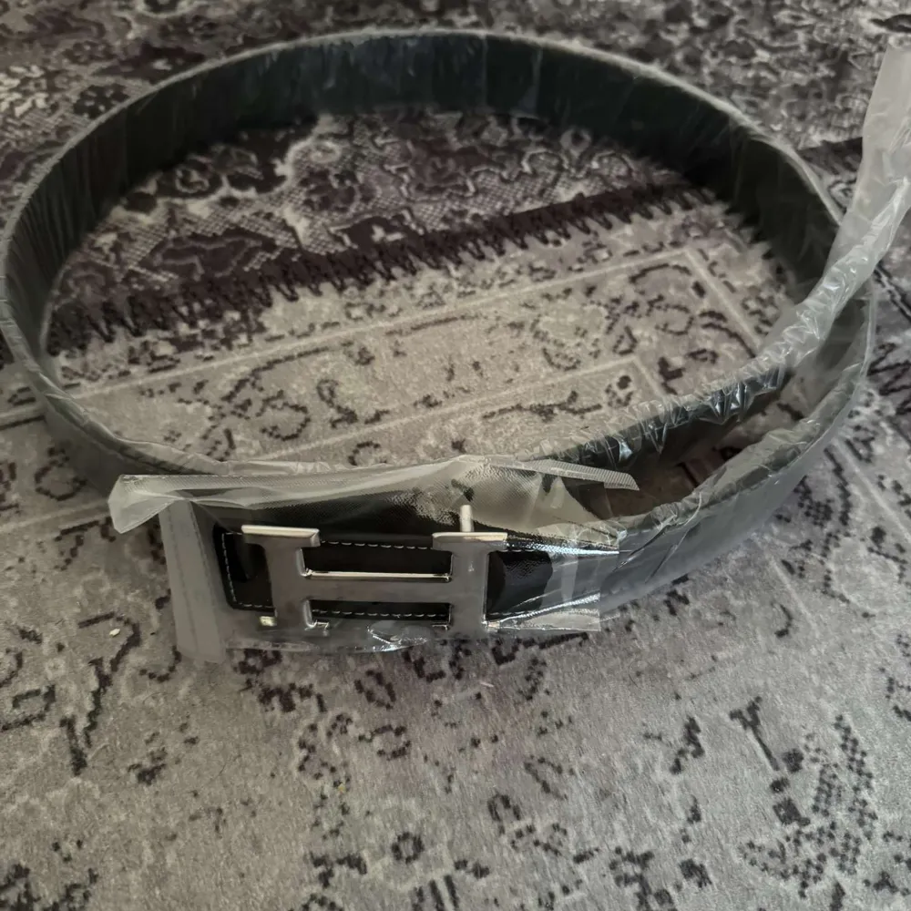 Snyggt svart bälte med ett stort silverfärgat H-format spänne. Perfekt för att ge en stilren touch till din outfit. Bältet är tillverkat i ett elegant material och har en klassisk design.. Muu.