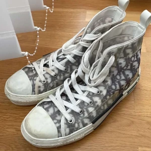 Mönstrade sneakers från Dior - Snygga höga sneakers från Dior med ett unikt mönster i grått och vitt. Skorna har snörning och en robust sula med Dior-logga på sidan. Perfekta för att ge din outfit en lyxig touch.