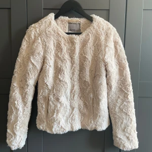 Beige pälsjacka (fuskpäls) - Beige pälsjacka i fuskpäls från Vero Moda. Knappar framtill och en ficka per sida. Kort modell.
