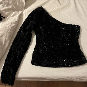Svart paljettopp från Bik Bok - Säljer en glittrig svart paljettopp från Bik Bok i storlek XS. Toppen har en one-shoulder design med en lång ärm och är perfekt för festliga tillfällen. Den är täckt av glänsande paljetter som ger en lyxig känsla. Köp direkt eller ge prisförslag! Skulle också säga att den är i mycket bättre kvalitet än andra paljett toppar jag har paljetterna kommer inte lossna på denna 