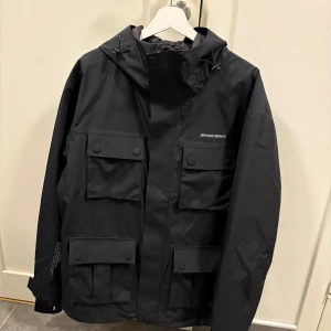 Svart jacka från Beyond Medals - Säljer en svart jacka från Beyond Medals med flera praktiska fickor och en justerbar huva. Jackan har en stilren design med knappar och dragkedja framtill. Perfekt för både stad och natur. Priset kan diskuteras.