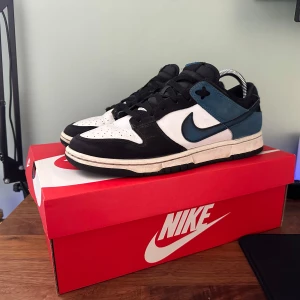 Nike Dunk - Snygga Nike Dunk sneakers i svart och vit med blå detaljer. Skorna har en klassisk design med snörning och rund tå. Perfekta för en stilren look.