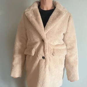 Beige pälsjacka - Säljer en mysig beige pälsjacka med långa ärmar och knappar framtill. Jackan har en klassisk krage och två fickor på sidorna. Perfekt för kyliga dagar. Barn storlek, men sitter som en S