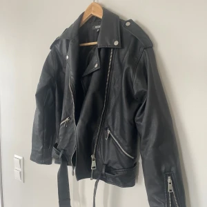 Svart skinnjacka från Missguided - Snygg svart skinnjacka från Missguided med klassisk bikerstil, silvriga dragkedjor, bred krage och bälte i midjan. Jackan har långa ärmar och coola detaljer som ger en edgy vibe till din outfit.